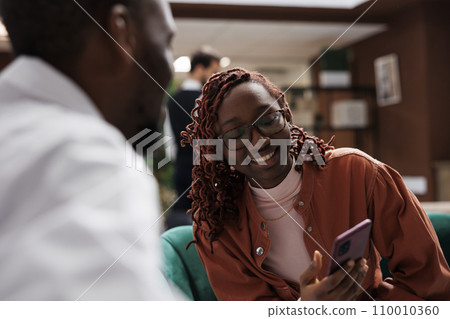 Hotel guests waiting to do check in, relaxing on couch in lounge area with smartphone. Man and woman sitting at reception lobby with marble floor, preparing to fill in registration files. Hotel guests waiting to do check in, relaxing on couch in lounge area with smartphone. Man and woman sitting at reception lobby with marble floor, preparing to fill in registration files. 110010360