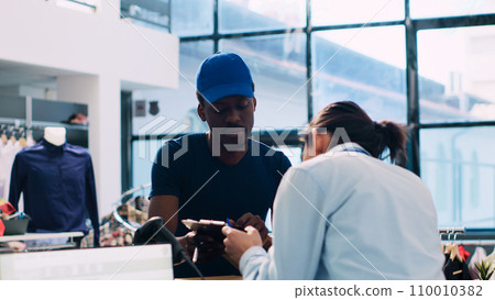 Asian employee signing distribution report, discussing delivery details with african american courier in shopping mall. Manager preparing online orders for shipping in modern boutique 110010382