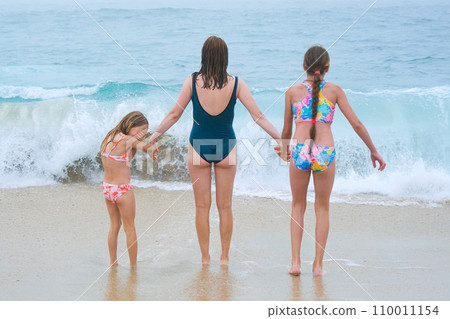 Mother and children playing on the ocean beach. Family enjoying the ocean. Mother holds girls's hands and they all look at the ocean together. Mother and children playing on the ocean beach. Family enjoying the ocean. Mother holds girls's hands and they all look at the ocean together. 110011154