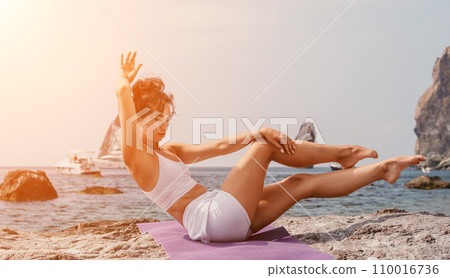 Fitness woman sea. A happy middle aged woman in white sportswear exercises morning outdoors on a beach with volcanic rocks by the sea. Female fitness pilates yoga routine concept. Healthy lifestyle. 110016736
