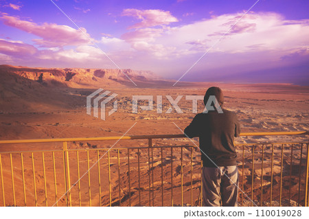 Young man standing on a viewpoint on the mount (Masada) and gazing sunrise in the desert. The man looking at the valley. Dead sea region, Masada, Israel 110019028