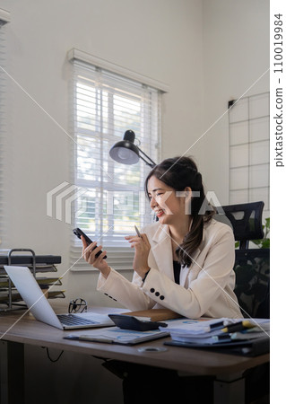 Young business woman on the phone at office. Business woman texting on the phone and working on laptop. Pretty young business woman sitting on workplace Young business woman on the phone at office. Business woman texting on the phone and working on laptop. Pretty young business woman sitting on workplace 110019984