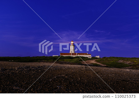 Panoramic view of cape Espichel lighthouse, Portugal, during sunset hours. Clear sky, few clouds, lighthouse sending light beams for safe maritime navigation, little foliage of rugged terrain.  110025373