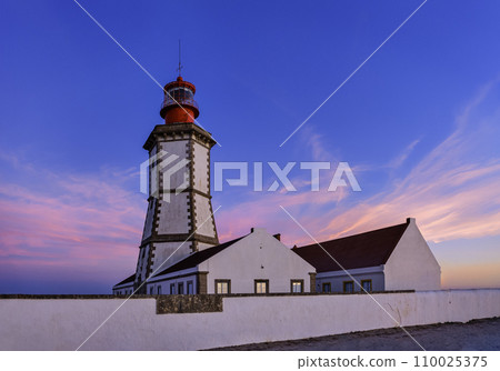Beautiful view of cape Espichel lighthouse in Portugal during colorful sunset. Traditional Portuguese architecture, clear sky, clouds, whitewashed walls and fence, summer landscape.  110025375
