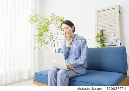 A middle-aged woman wearing pajamas and using a laptop while yawning at home 110027754