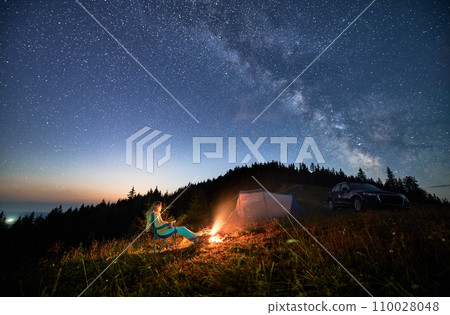 Night camping in the mountains under starry sky full of stars. Travelling woman admiring starry night in mountains. Young woman sitting next to camp fire, tourist tent and off-road car. 110028048