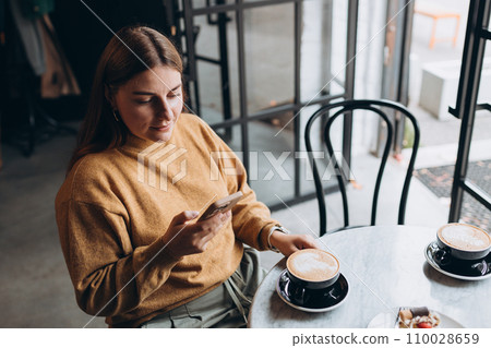 Beautiful young woman in casual clothes smiling, drinking a cup of coffee and holding a phone in her hands while spending time in the cafe. Modern interior. Food concept 110028659