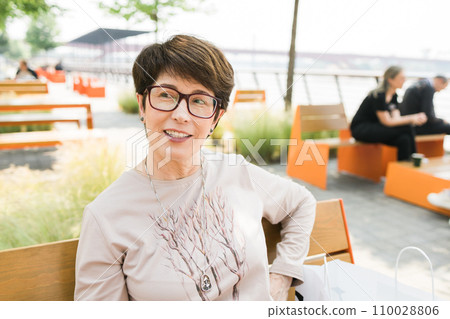 Outdoor portrait of beautiful and elegant middle age 55 - 60 year old woman wearing long sleeve top sitting on bench on the street. Copy space and empty place for text advertising 110028806