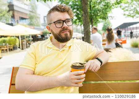 Millennial smiling student man in yellow polo t-shirt eyeglasses sitting on bench drink coffee have breakfast rest relax in sunshine spring green city park outdoors on nature. Urban leisure concept. 110029186