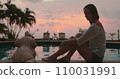 Woman relaxes sitting near pool on tropical island hotel resort. Bright colorful sunset sky and coconut palms reflected in water. Travel, tourism, holiday spa vacation. 110031991
