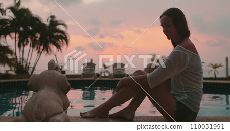 Woman relaxes sitting near pool on tropical island hotel resort. Bright colorful sunset sky and coconut palms reflected in water. Travel, tourism, holiday spa vacation. Woman relaxes sitting near pool on tropical island hotel resort. Bright colorful sunset sky and coconut palms reflected in water. Travel, tourism, holiday spa vacation. 110031991