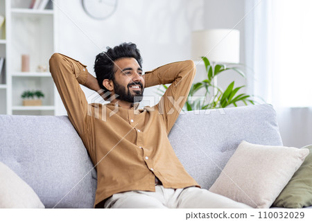 Happy Indian young man sitting relaxed on sofa at home with hands behind head and resting, smiling contentedly. 110032029