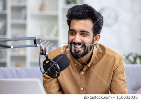 Close-up photo of a young Indian man smiling sitting on the couch at home in front of a microphone stand and talking. 110032030