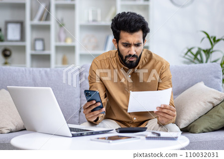 Worried young Indian man sitting on sofa at home, working on laptop, looking sadly at documents and bills, holding phone. 110032031