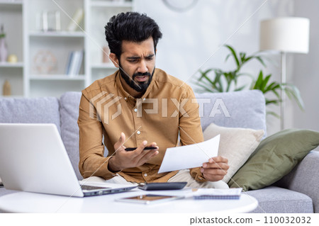 Indian young man working from home, sitting on sofa and checking bills and documents, crumpling papers in hands, looking at data frustrated and upset. 110032032