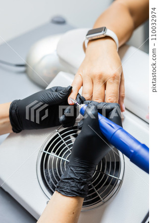 Manicurist uses an electric drill to remove old gel polish from nails. A woman is getting a manicure of nails. The beautician files the client's nails. professional manicure tool. Close-up 110032974