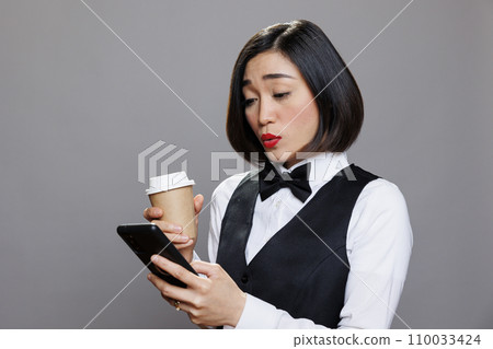 Asian woman in waitress uniform sending message on mobile phone during coffee break. Young receptionist talking on mobile phone social media and drinking hot beverage to go Asian woman in waitress uniform sending message on mobile phone during coffee break. Young receptionist talking on mobile phone social media and drinking hot beverage to go 110033424