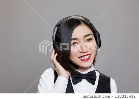 Smiling asian waitress in uniform listening to music in wireless headphones. Cheerful young attractive restaurant receptionist wearing earphones and enjoying playlist while posing in studio 110033432