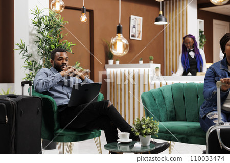 Tired African American man entrepreneur traveling on business, sitting with laptop in hotel lobby checking email. Black guy freelancer working remotely while waiting for check-in 110033474