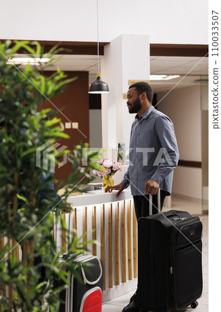 Young African American man with suitcase standing at reception area, talking pleasantly with front desk employees, waiting in line to check-in after arrival. Black guy tourist checking out of hotel Young African American man with suitcase standing at reception area, talking pleasantly with front desk employees, waiting in line to check-in after arrival. Black guy tourist checking out of hotel 110033507