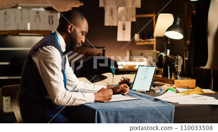 Couturier drawing sketches clothes at workstation, preparing to design new clothing line handmade. Fashion designer working on modern items, looking at models on tablet in atelier. Couturier drawing sketches clothes at workstation, preparing to design new clothing line handmade. Fashion designer working on modern items, looking at models on tablet in atelier. 110033508