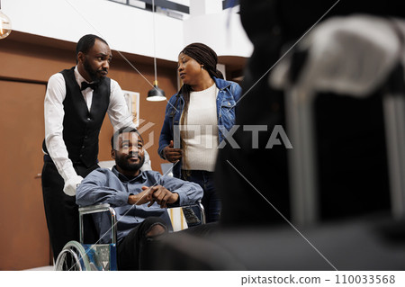 Hotel bellman transferring disabled guest on wheelchair to room, happy young African American handicapped man enjoying customer service for people with disabilities, accessible tourism Hotel bellman transferring disabled guest on wheelchair to room, happy young African American handicapped man enjoying customer service for people with disabilities, accessible tourism 110033568