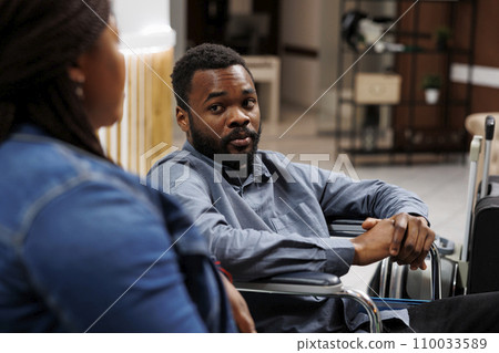 African American man hotel guest with reduced mobility sitting in lobby with wife and waiting for check-in procedure, traveling with disabilities. Wheelchair accessible accommodations 110033589