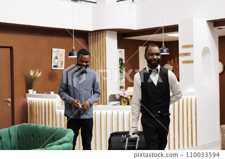 Smiling friendly hotel bellboy helping guest with his luggage after check-in procedure, african american guy bellhop pulling suitcase delivering baggage to room. Bell desk staff Smiling friendly hotel bellboy helping guest with his luggage after check-in procedure, african american guy bellhop pulling suitcase delivering baggage to room. Bell desk staff 110033594