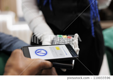 Waiter holding POS terminal accepting cashless transaction from customer in restaurant or hotel. Client paying with phone, making mobile payment, using NFC payment app, close up 110033596