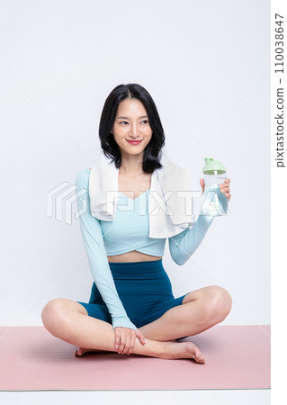 There is a woman sitting on a yoga mat with a water bottle in one hand and a towel wrapped around her shoulder. There is a woman sitting on a yoga mat with a water bottle in one hand and a towel wrapped around her shoulder. 110038647