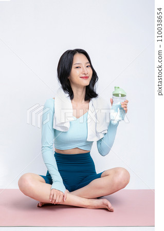 There is a woman sitting on a yoga mat with a water bottle. There is a woman sitting on a yoga mat with a water bottle. 110038654