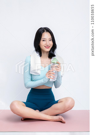 There is a woman sitting with a towel on a yoga mat with a water bottle in her hand There is a woman sitting with a towel on a yoga mat with a water bottle in her hand 110038655