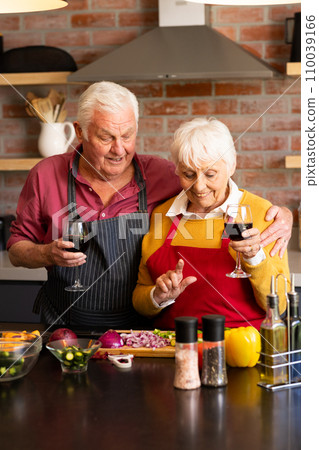 Happy caucasian senior couple preparing vegetables, drinking wine, embracing in kitchen, copy space 110039166