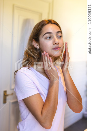 Happy biracial woman touching face looking in bathroom mirror and smiling at home 110039331