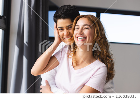 Portrait of happy biracial lesbian couple embracing in sun by window at home 110039333