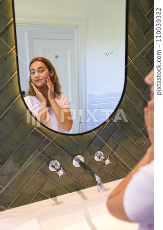 Happy biracial woman touching face and smiling, looking at reflection in bathroom mirror at home Happy biracial woman touching face and smiling, looking at reflection in bathroom mirror at home 110039382