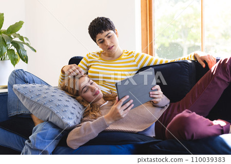 Happy biracial lesbian couple relaxing on couch together using tablet at home 110039383