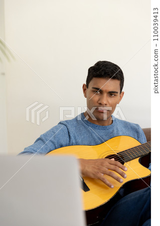 Biracial man sitting on sofa playing guitar and using laptop at home 110039413