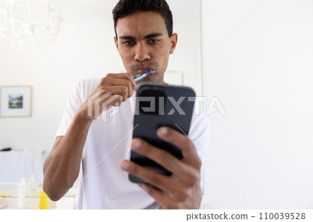 Biracial man brushing teeth and using smartphone in bathroom at home 110039528