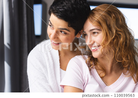 Happy biracial lesbian couple embracing in sun looking out of window at home 110039563
