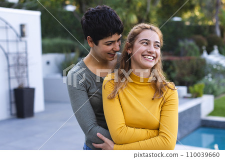 Happy biracial lesbian couple standing on garden terrace by pool embracing and smiling 110039660