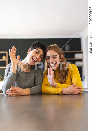 Happy biracial lesbian couple sitting at dining table having video call at home, waving, copy space Happy biracial lesbian couple sitting at dining table having video call at home, waving, copy space 110039684