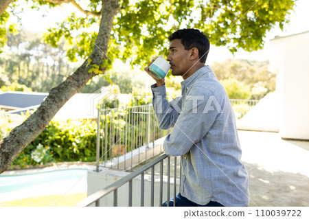 Thoughtful biracial man drinking coffee and standing on balcony at home, copy space 110039722