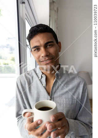 Portrait of happy biracial man holding mug next to window at home 110039723