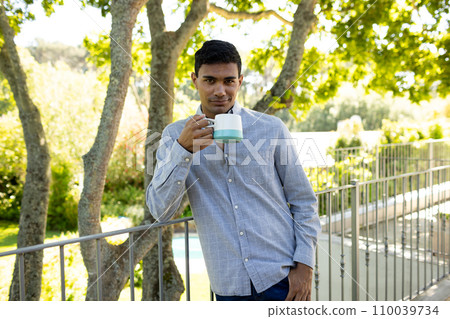 Portrait of happy biracial man holding mug on balcony at home Portrait of happy biracial man holding mug on balcony at home 110039734
