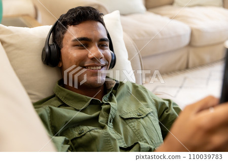 Happy biracial man wearing headphones lying on sofa using tablet at home Happy biracial man wearing headphones lying on sofa using tablet at home 110039783