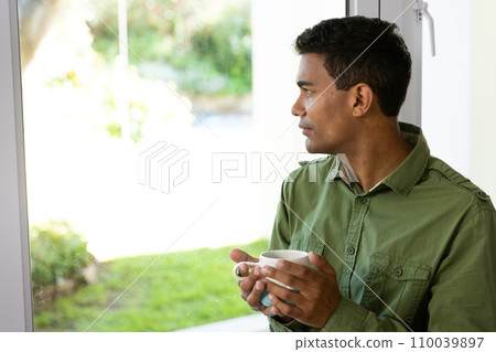 Thoughtful biracial man holding mug looking through window at home Thoughtful biracial man holding mug looking through window at home 110039897