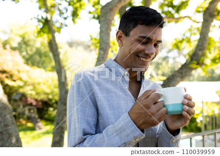 Happy biracial man drinking coffee and standing on balcony at home 110039959