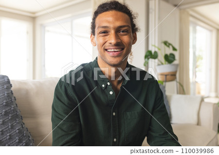 Happy biracial man with long curly hair having video call and smiling in sunny living room at home 110039986