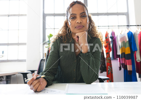 Happy biracial female fashion designer having video call and taking notes at desk in studio 110039987